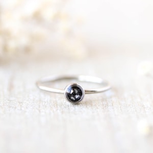 May include: A delicate silver ring with a small, dark, round stone set in the center. The ring band is thin and polished, and the stone appears to have a speckled or textured surface. The background is softly blurred with white and cream tones.