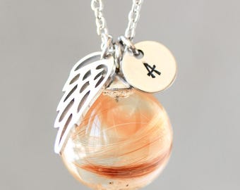 May include: A silver necklace with a clear orb pendant containing strands of brown and orange hair. The pendant is adorned with a silver wing charm and a small circular charm with the letter "A". The necklace hangs against a gray background.