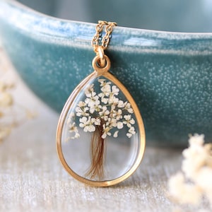 Op de afbeelding: Een goudkleurige druppelvormige hangerketting. De hanger bevat een miniatuurboom gemaakt van witte bloemen en bruine strengen, ingekapseld in heldere hars. De ketting is ook goudkleurig.