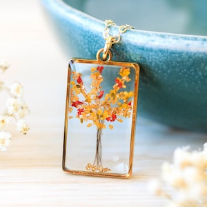 Puede incluir: Un collar con colgante dorado con un colgante rectangular de resina transparente. Dentro del colgante hay un diseño de árbol en miniatura con flores amarillas y rojas. El collar está sobre un fondo borroso con un cuenco turquesa y flores blancas.