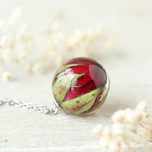 Op de afbeelding: Een bolvormige hanger aan een ketting met een geconserveerde dieprode rozenknop ingesloten in heldere hars. De hanger is bevestigd aan een dunne zilverkleurige metalen ketting en toont de natuurlijke groene kelkbladeren en rode bloemblaadjes.