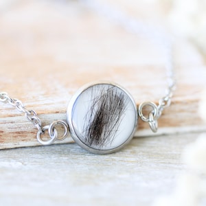Op de afbeelding: Een zilveren armband met een ronde hanger met een bundel donker haar tegen een witte achtergrond. De hanger is omlijst in zilver, en de delicate ketting is zichtbaar. De achtergrond toont een lichtgekleurd houten oppervlak.