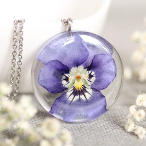 Puede incluir: Un collar con colgante redondo y transparente con una flor de pensamiento morada prensada. La flor está encapsulada en resina y suspendida de una cadena plateada. El colgante se muestra sobre una superficie gris con flores blancas en el fondo.