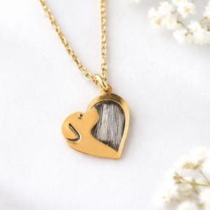 Puede incluir: Un collar con colgante en forma de corazón dorado con una silueta de perro y una sección llena de pelo gris. El collar cuelga de una delicada cadena dorada. Flores blancas en el fondo.