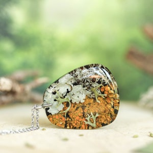 Flat-lying view of a triangular botanical resin pendant filled with vibrant orange and white lichens. The piece is placed horizontally on a neutral background with hints of forest elements