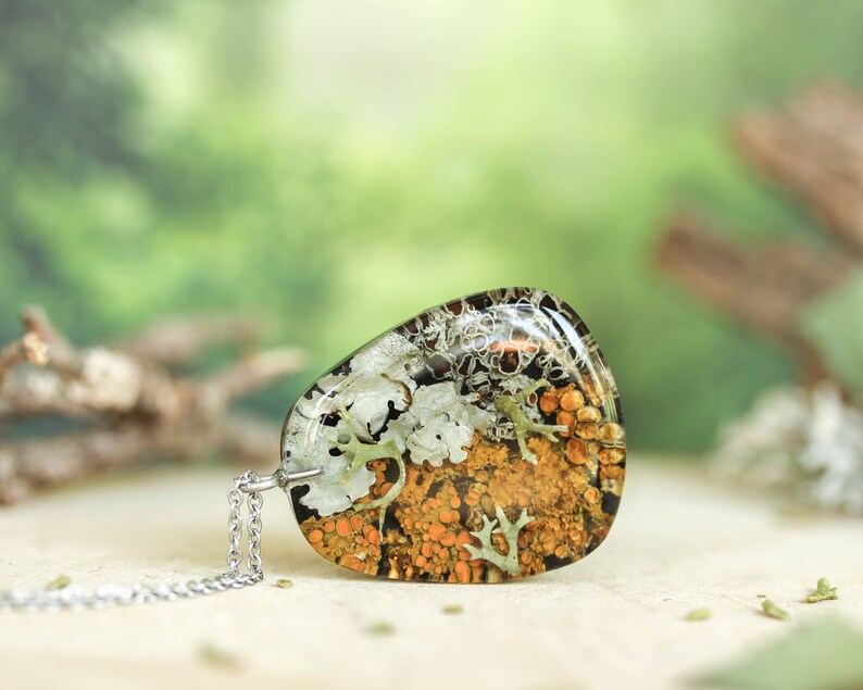 Flat-lying view of a triangular botanical resin pendant filled with vibrant orange and white lichens. The piece is placed horizontally on a neutral background with hints of forest elements
