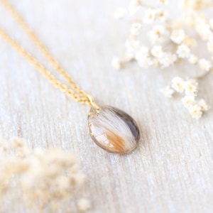 Pode incluir: Um colar de corrente dourada com um pingente em forma de gota. O pingente cont&eacute;m uma mistura de materiais brancos, castanhos e dourados. O colar &eacute; exibido em uma superf&iacute;cie de madeira clara, com flores brancas ao fundo.