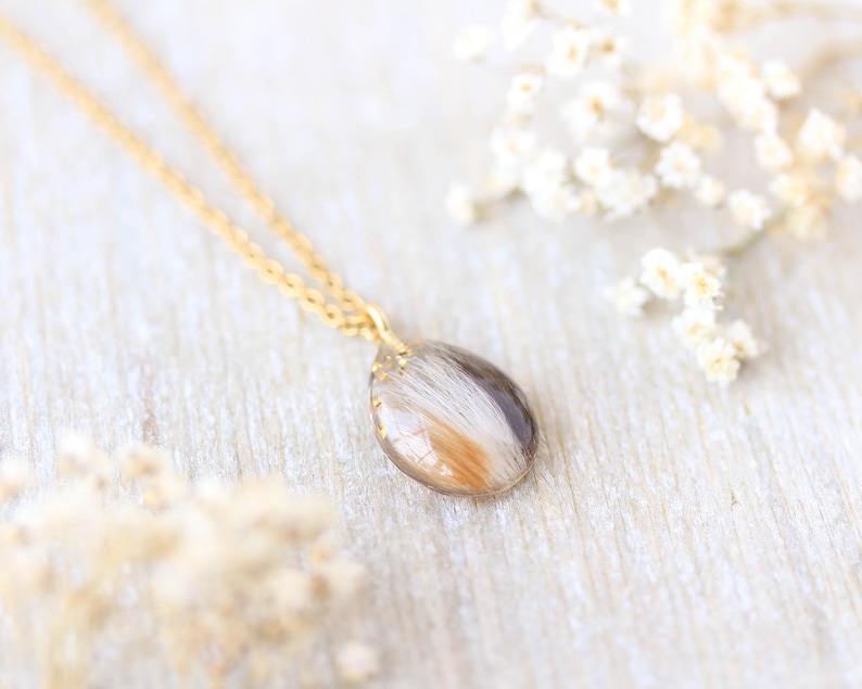 Pode incluir: Um colar de corrente dourada com um pingente em forma de gota. O pingente cont&eacute;m uma mistura de materiais brancos, castanhos e dourados. O colar &eacute; exibido em uma superf&iacute;cie de madeira clara, com flores brancas ao fundo.