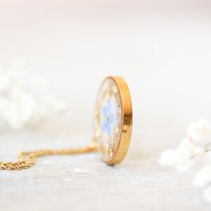 Puede incluir: Un collar con colgante dorado con un peque&ntilde;o dije redondo de resina transparente. El dije contiene una flor azul y copos dorados. La cadena del collar tambi&eacute;n es dorada. El fondo est&aacute; borroso con flores blancas.