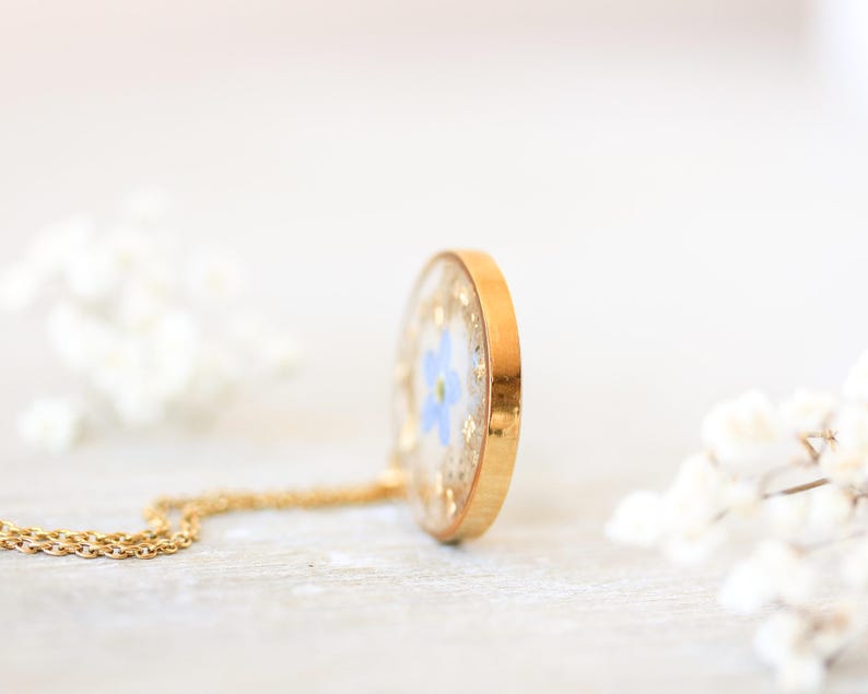 Puede incluir: Un collar con colgante dorado con un peque&ntilde;o dije redondo de resina transparente. El dije contiene una flor azul y copos dorados. La cadena del collar tambi&eacute;n es dorada. El fondo est&aacute; borroso con flores blancas.