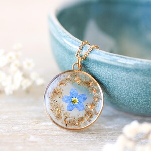 Puede incluir: Un collar con colgante dorado con un dije redondo transparente de resina. El dije contiene una peque&ntilde;a flor azul y copos dorados. El collar se muestra cerca de un cuenco turquesa y flores blancas.