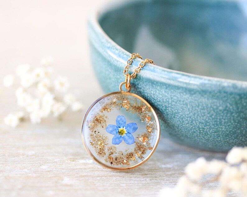 Puede incluir: Un collar con colgante dorado con un dije redondo transparente de resina. El dije contiene una peque&ntilde;a flor azul y copos dorados. El collar se muestra cerca de un cuenco turquesa y flores blancas.