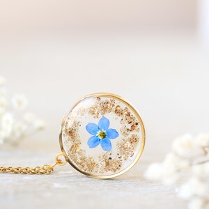 Puede incluir: Un collar con colgante dorado con un dije redondo y transparente de resina. Dentro del dije hay una peque&ntilde;a flor azul nomeolvides con copos dorados. La cadena del collar tambi&eacute;n es dorada. El fondo es de un color suave y neutro.