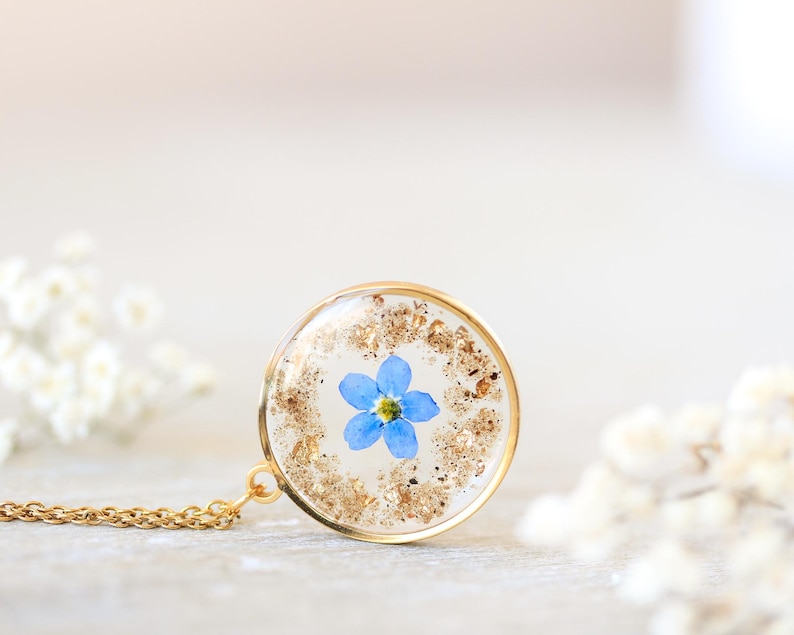 Puede incluir: Un collar con colgante dorado con un dije redondo y transparente de resina. Dentro del dije hay una peque&ntilde;a flor azul nomeolvides con copos dorados. La cadena del collar tambi&eacute;n es dorada. El fondo es de un color suave y neutro.