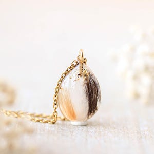 Pode incluir: Um colar de corrente dourada com um pingente em forma de gota. O pingente &eacute; transparente e cont&eacute;m mechas de cabelo castanho, bege e branco. O colar &eacute; exibido em uma superf&iacute;cie clara.