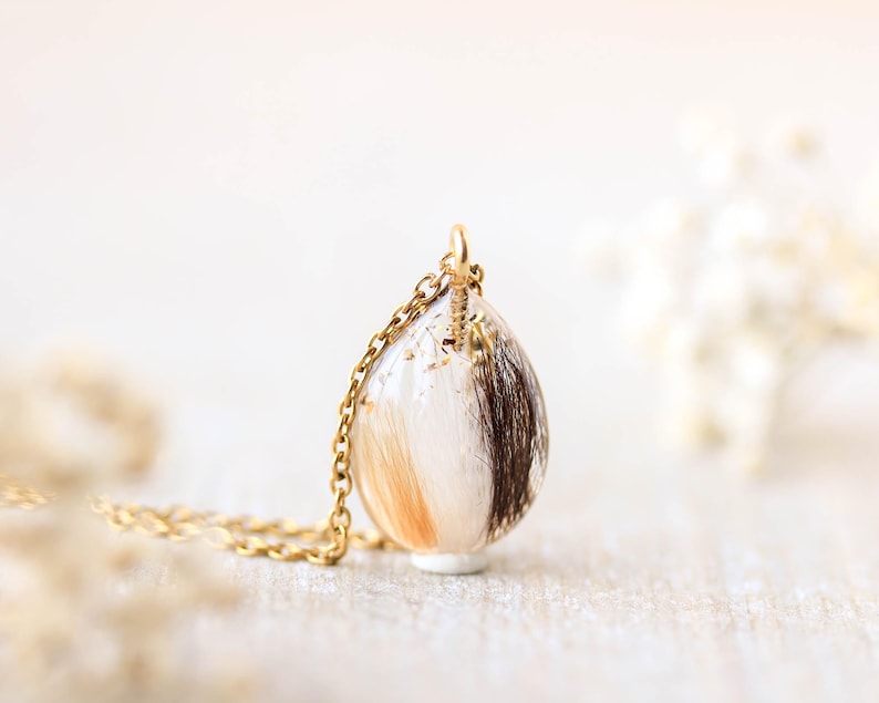 Pode incluir: Um colar de corrente dourada com um pingente em forma de gota. O pingente &eacute; transparente e cont&eacute;m mechas de cabelo castanho, bege e branco. O colar &eacute; exibido em uma superf&iacute;cie clara.