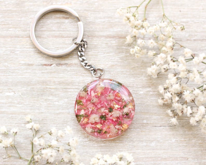 May include: A circular keyring pendant featuring a pink floral design set in clear resin. The pendant is connected to a silver chain and ring. White flowers are arranged around the keyring on a light wood surface.
