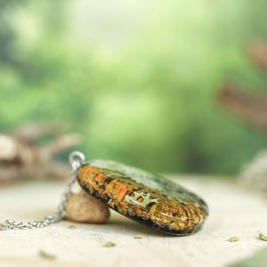 Side view of a handmade botanical resin necklace showcasing embedded orange and white lichens inside a translucent pendant. The piece rests on a small stone with a nature-inspired background