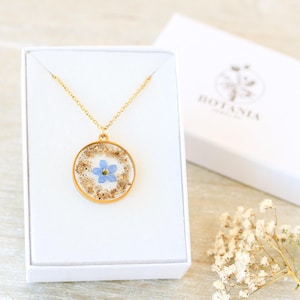 Puede incluir: Un collar dorado con un colgante circular que contiene una peque&ntilde;a flor azul y motas doradas. El colgante se muestra en una caja blanca. La caja tiene la palabra "BOTANIA" impresa en la tapa.