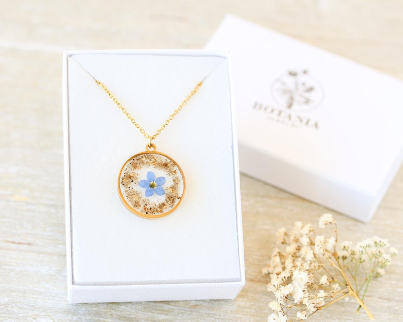 Puede incluir: Un collar dorado con un colgante circular que contiene una peque&ntilde;a flor azul y motas doradas. El colgante se muestra en una caja blanca. La caja tiene la palabra "BOTANIA" impresa en la tapa.