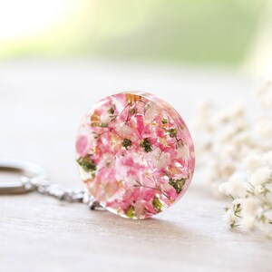 May include: A circular keychain pendant made of clear resin, containing pink and white dried flowers and green leaves. It's attached to a silver-tone chain and keyring. The background is blurred, with white flowers and greenery.