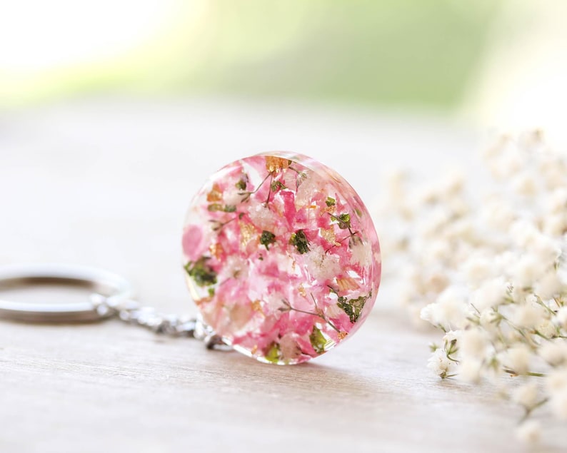 May include: A circular keychain pendant made of clear resin, containing pink and white dried flowers and green leaves. It's attached to a silver-tone chain and keyring. The background is blurred, with white flowers and greenery.