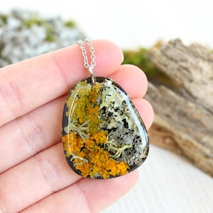 Close-up of a hand holding a nature-inspired botanical resin pendant with embedded real lichens in vibrant orange and pale gray hues. The pendant has a soft rounded triangle shape and a silver chain