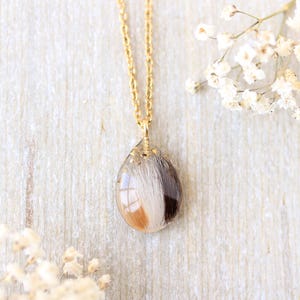 Pode incluir: Um colar de corrente dourada com um pingente em forma de gota. O pingente transparente cont&eacute;m uma mistura de pelo branco, castanho e bege. O colar &eacute; exibido sobre uma superf&iacute;cie de madeira clara, com flores brancas ao fundo.