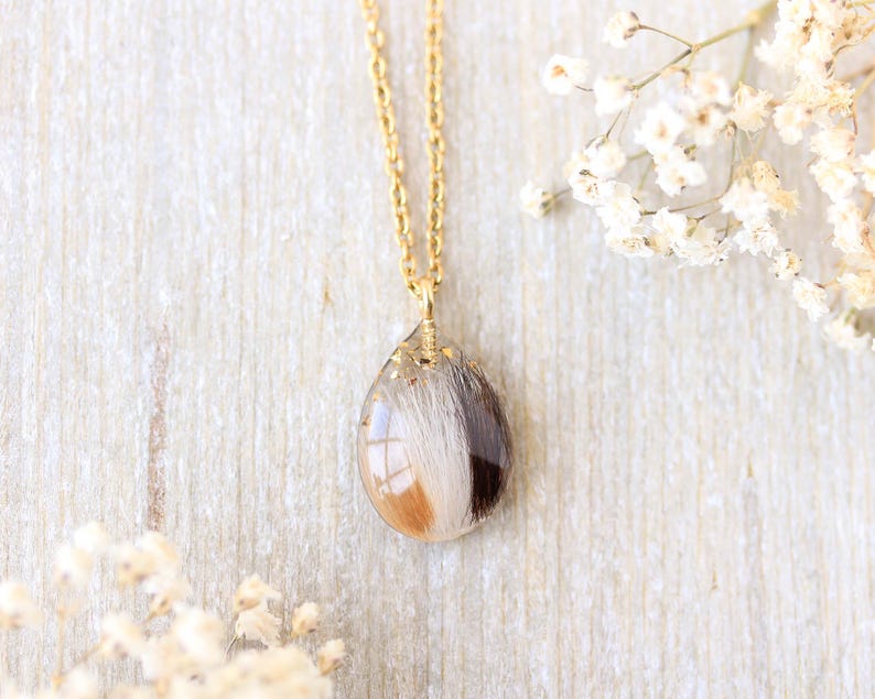 Pode incluir: Um colar de corrente dourada com um pingente em forma de gota. O pingente transparente cont&eacute;m uma mistura de pelo branco, castanho e bege. O colar &eacute; exibido sobre uma superf&iacute;cie de madeira clara, com flores brancas ao fundo.