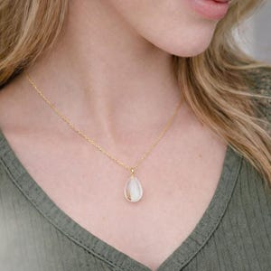 Pode incluir: Um colar dourado com um pingente em forma de gota. O pingente tem um centro branco perolado. O colar &eacute; usado sobre uma camisa verde.