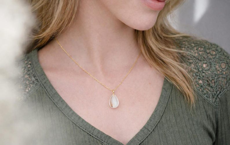 Pode incluir: Um colar dourado com um pingente em forma de gota. O pingente tem um centro branco perolado. O colar &eacute; usado sobre uma camisa verde.