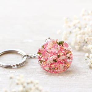 May include: A circular resin pendant keychain, filled with pink flowers and gold leaf, attached to a silver keyring. The background features white flowers. The pendant's diameter is approximately 3 cm.