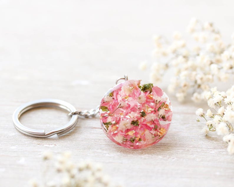 May include: A circular resin pendant keychain, filled with pink flowers and gold leaf, attached to a silver keyring. The background features white flowers. The pendant's diameter is approximately 3 cm.