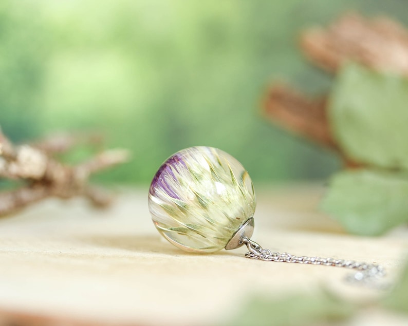 May include: A silver chain necklace with a clear glass pendant containing a preserved flower with purple and green hues.
