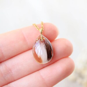 Pode incluir: Colar com corrente dourada e pingente em forma de gota. O pingente &eacute; feito de resina transparente, contendo uma mistura de cabelo castanho, branco e castanho claro. O colar &eacute; segurado em uma m&atilde;o.