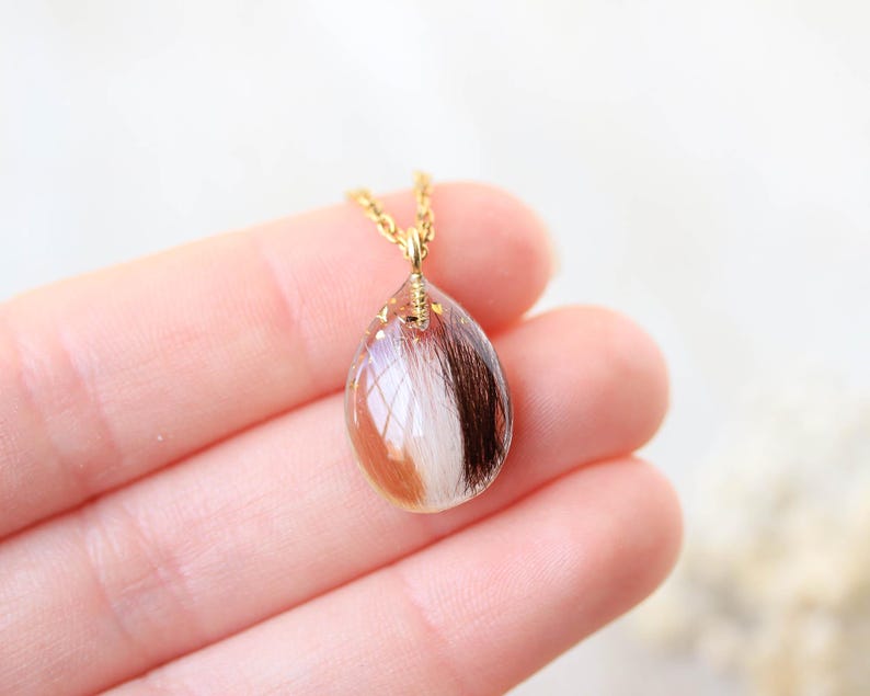Pode incluir: Colar com corrente dourada e pingente em forma de gota. O pingente &eacute; feito de resina transparente, contendo uma mistura de cabelo castanho, branco e castanho claro. O colar &eacute; segurado em uma m&atilde;o.