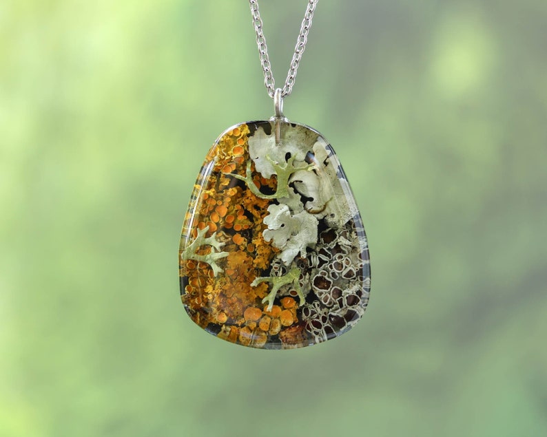 Pendant necklace crafted with real preserved lichens in earthy orange, yellow, and soft gray tones. The triangular shape highlights the organic composition, with a silver chain and blurred forest background