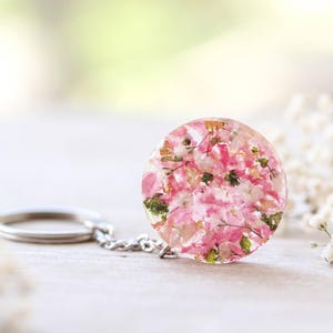 May include: A circular keychain pendant made of clear resin, containing pink and white dried flowers and gold leaf. It is attached to a silver chain and keyring. The background is blurred, with white flowers and a soft, neutral colour palette.