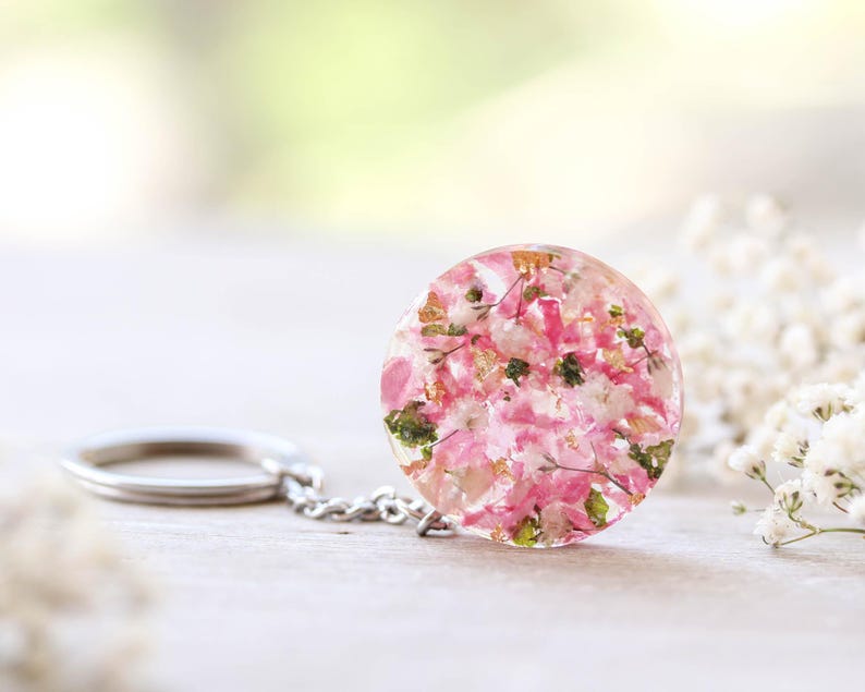May include: A circular keychain pendant made of clear resin, containing pink and white dried flowers and gold leaf. It is attached to a silver chain and keyring. The background is blurred, with white flowers and a soft, neutral colour palette.