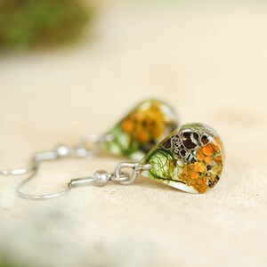 Close-up of a pair of teardrop-shaped earrings. The earrings feature resin accents with botanical elements in shades of orange, green, and gray. The earrings hang from delicate, silver-toned hooks.