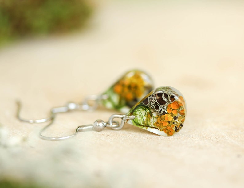 Close-up of a pair of teardrop-shaped earrings. The earrings feature resin accents with botanical elements in shades of orange, green, and gray. The earrings hang from delicate, silver-toned hooks.