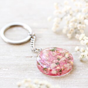 May include: A circular keyring with a silver chain and ring. The pendant is a clear resin circle filled with pink and white dried flowers and gold flakes. The background is a light wooden surface with white flowers.