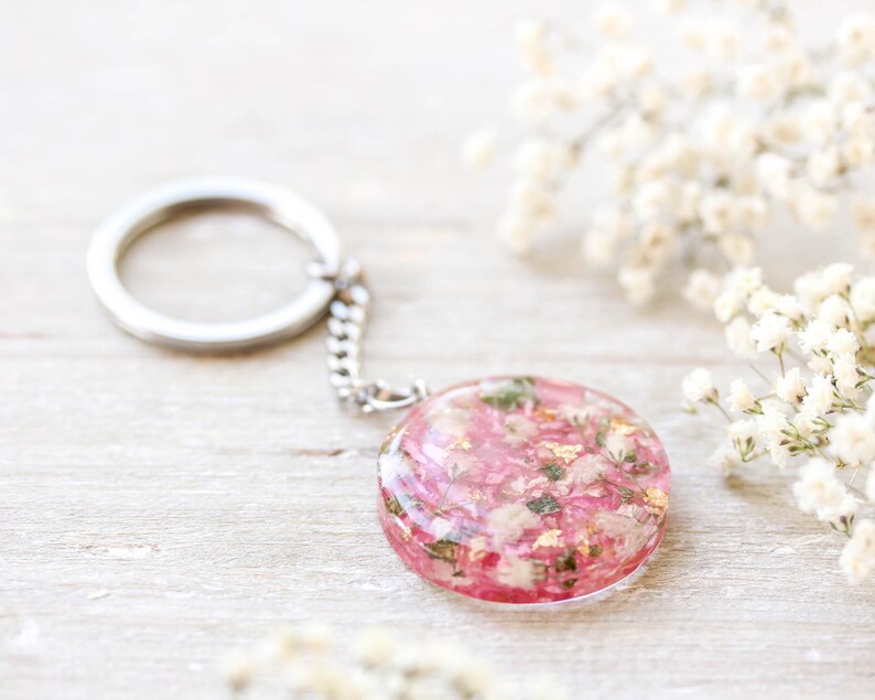 May include: A circular keyring with a silver chain and ring. The pendant is a clear resin circle filled with pink and white dried flowers and gold flakes. The background is a light wooden surface with white flowers.