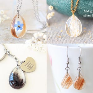 Pode incluir: Uma cole&ccedil;&atilde;o de pe&ccedil;as de joalharia em resina artesanais. Inclui um colar de corrente prateada com um pendente em forma de gota com uma flor azul e cabelo, um colar de corrente dourada com flocos dourados, um porta-chaves e brincos.