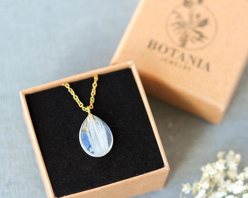 Pode incluir: Um colar com pingente transparente em forma de gota com detalhes brancos, apresentado em uma caixa forrada de veludo preto. O colar tem uma corrente dourada. A caixa est&aacute; dentro de uma caixa de papel&atilde;o marrom com o texto "BOTANIA JEWELRY".