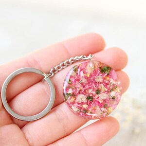 May include: A circular keyring pendant featuring pink and white flowers and gold leaf inclusions, set in clear resin. It's connected to a silver chain and a silver key ring.