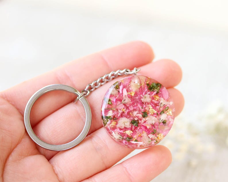 May include: A circular keyring pendant featuring pink and white flowers and gold leaf inclusions, set in clear resin. It's connected to a silver chain and a silver key ring.