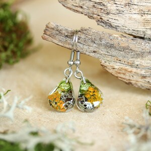 Teardrop-shaped resin earrings with preserved orange and gray lichen and green moss. Resting on a beige surface beside driftwood and moss, photographed in soft daylight with a natural green blurred background.