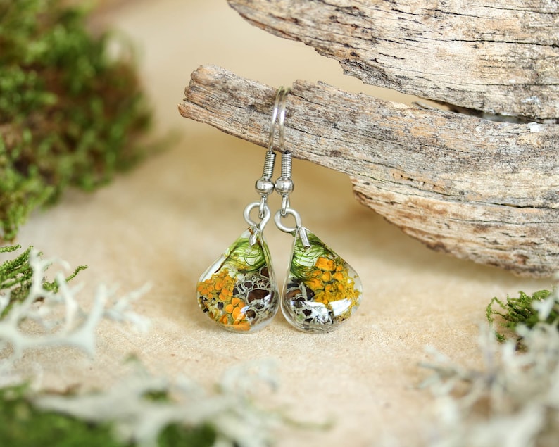 Teardrop-shaped resin earrings with preserved orange and gray lichen and green moss. Resting on a beige surface beside driftwood and moss, photographed in soft daylight with a natural green blurred background.