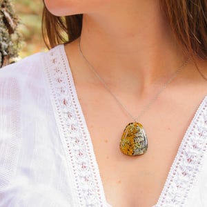 Model wearing a botanical resin necklace with real lichens in a warm-toned pendant. The piece stands out against a white embroidered top and natural outdoor background, showcasing its wearable scale